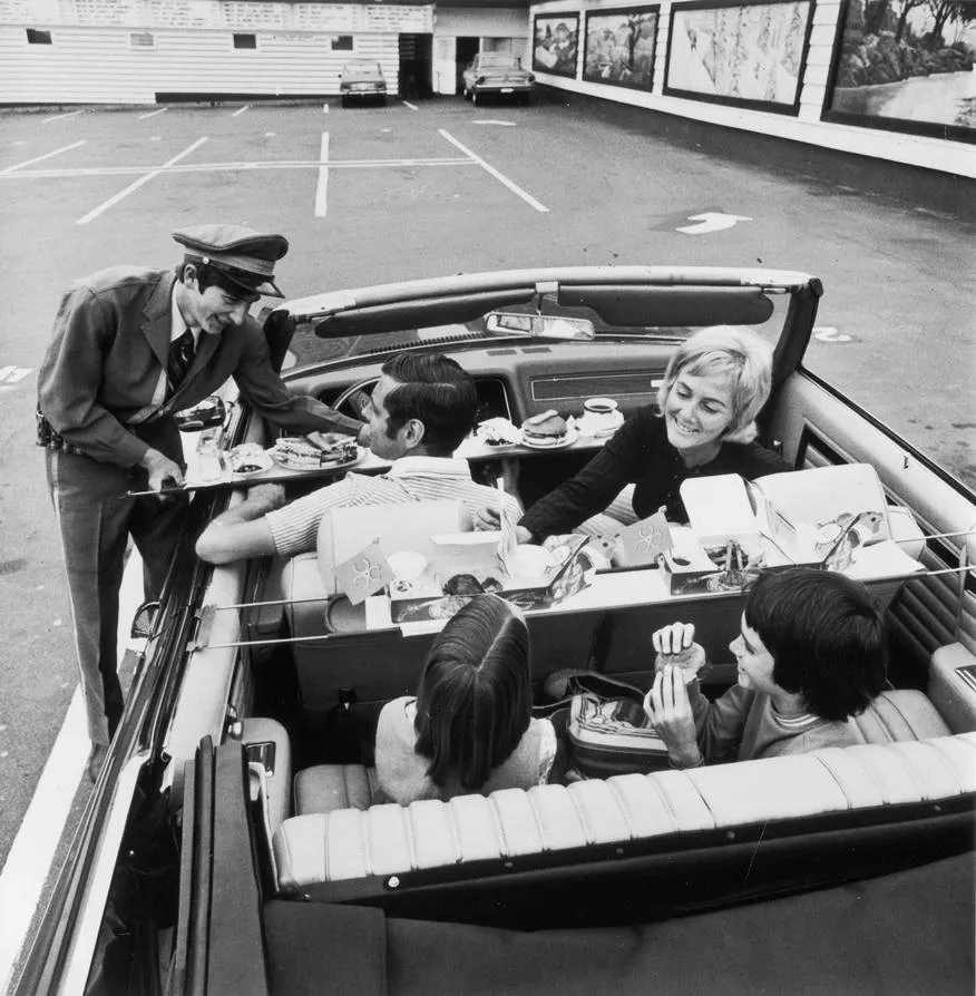 Black and white photograph of a White Spot car-hop in a peaked cap and uniform passing a tray of food through the window of a convertible, where a family of four sits with Pirate Pak boxes open on the seats, in a parking lot lined with painted murals