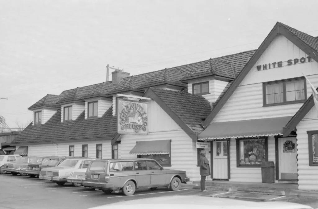 Black and white photograph of the original White Spot on Granville Street: a long white clapboard building with a steep shingled roof and dormer windows, a Granville House sign, striped awnings, and a row of 1970s cars parked out front